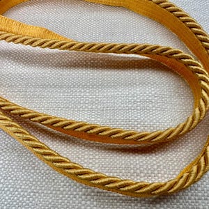 May include: A close-up of a gold braided trim with a rope-like texture. The trim is on a white fabric background.
