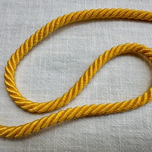 May include: A bright yellow twisted cord trim featuring a multi-strand rope construction with a smooth, lustrous finish. The cord is arranged in a curved, flowing shape, showcasing the consistent spiral texture and vibrant golden-yellow color.