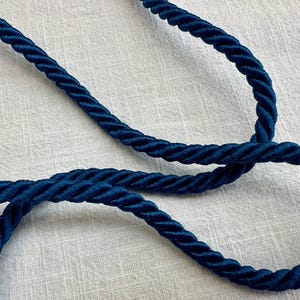 May include: A dark blue twisted cord made of multiple strands of fiber wound together. The cord features a smooth, slightly reflective texture and is arranged in a loose, overlapping loop pattern, showcasing the consistent thickness of the rope.