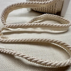 May include: A close-up of a cream-colored rope trim with a braided design. The trim is coiled and lying on a textured fabric surface.