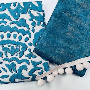 May include: Two pieces of teal fabric with a white pom-pom trim. One fabric has a white background with a teal pattern, and the other fabric is a solid teal color.