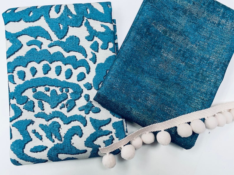 May include: Two pieces of teal fabric with a white pom-pom trim. One fabric has a white background with a teal pattern, and the other fabric is a solid teal color.