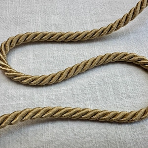 May include: A single length of gold-colored twisted cord trim. The cord features a continuous, spiraling rope-like texture with a metallic sheen, arranged in a gentle, winding curve across the frame to showcase its uniform thickness and weave.