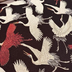 May include: Close-up of a fabric with a repeating pattern of white and red cranes against a dark brown background. The cranes are depicted in flight with detailed wing and feather designs. The fabric has a textured appearance.