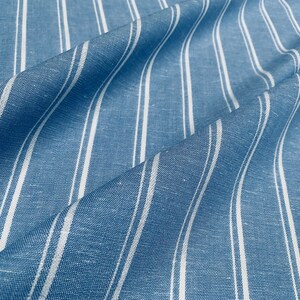 Pu&ograve; includere: Tessuto a righe blu e bianche con una texture di lino. Il tessuto &egrave; piegato e drappeggiato, mostrando il motivo e la texture.