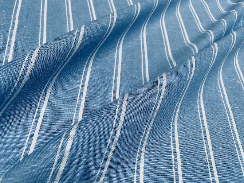 Pu&ograve; includere: Tessuto a righe blu e bianche con una texture di lino. Il tessuto &egrave; piegato e drappeggiato, mostrando il motivo e la texture.