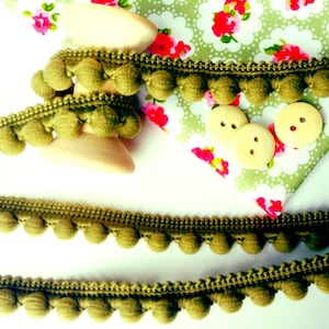May include: Olive green pom pom trim with wooden spool and three wooden buttons. The trim is on a white surface with a floral fabric in the background.