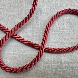 May include: A length of twisted rope trim in a deep dusty rose color. The cord features a tightly coiled, multi-strand construction with a subtle sheen, arranged in a loose, overlapping loop pattern that highlights the uniform texture.