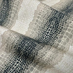 May include: Close-up of a fabric with a reptile skin pattern. The design features a mix of neutral colors, including beige, gray, and black, creating a textured and layered effect. The fabric is folded, showing the pattern's repeating scales.