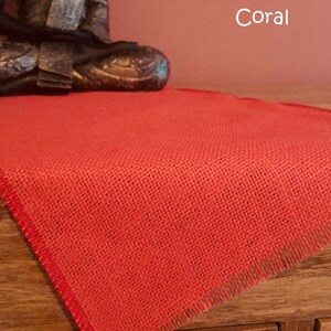 May include: Coral coloured burlap table runner with frayed edges. The runner is on a wooden surface.
