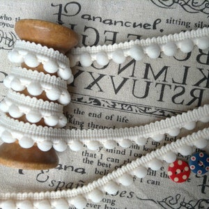 May include: White pom-pom trim wound around a wooden spool. The trim is a white cotton ribbon with small white pom-poms along the edge.