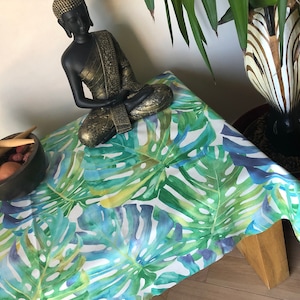 May include: A white tablecloth with a tropical leaf pattern in shades of green, blue, and yellow. A black Buddha statue sits on the tablecloth, and a bowl of maracas is in the foreground.