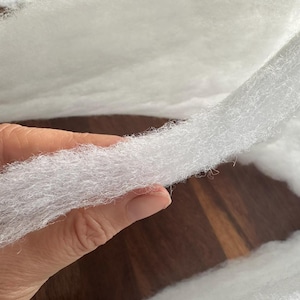 May include: Close-up of white, fluffy batting material. The batting is held in a hand, showing its soft, textured appearance. The material appears to be a thick, continuous sheet, ideal for crafts or quilting.