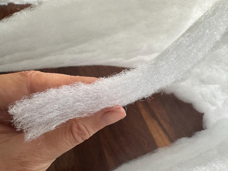 May include: Close-up of white, fluffy batting material. The batting is held in a hand, showing its soft, textured appearance. The material appears to be a thick, continuous sheet, ideal for crafts or quilting.