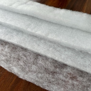 May include: Close-up of three layers of soft, white, and gray batting fabric. The fabric is layered diagonally, with the bottom layer being a darker gray and the top layer being a bright white. The fabric has a fluffy, textured appearance.