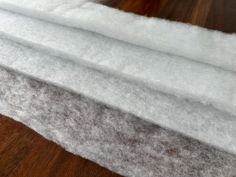 May include: Close-up of three layers of soft, white, and gray batting fabric. The fabric is layered diagonally, with the bottom layer being a darker gray and the top layer being a bright white. The fabric has a fluffy, textured appearance.