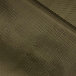 Op de afbeelding: Close-up van een getextureerd, olijfgroen oppervlak met een rasterpatroon. Het oppervlak lijkt te zijn gemaakt van hout of een vergelijkbaar materiaal, met diagonale lijnen die elkaar kruisen om een ​​gestructureerd effect te creëren. De belichting creëert schaduwen en highlights.