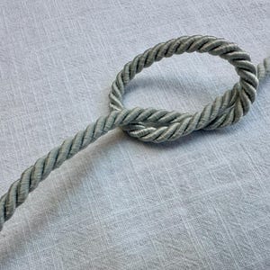May include: A single length of light grey twisted cord featuring a decorative knot tied in the centre. The cord has a smooth, uniform texture and a consistent thickness throughout its length, showcasing a classic braided construction style.