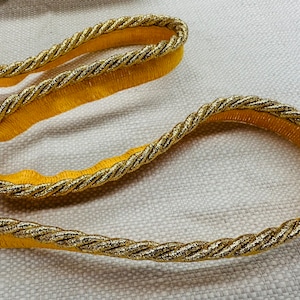 May include: Gold and yellow trim with a rope-like texture. The trim is made of a soft, woven fabric with a gold thread running through it.