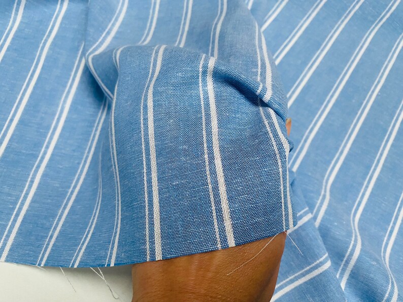Pu&ograve; includere: Tessuto a righe blu e bianche con una trama di lino. Il tessuto &egrave; piegato e drappeggiato su una mano.