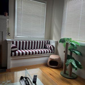 May include: A built-in window seat with a striped burgundy and white cushion and two matching bolster pillows. The room also features a cat bed, a cat tree, and a glass coffee table.