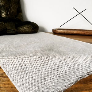 May include: A white linen table runner with frayed edges, placed on a wooden surface. A small wooden incense holder with two incense sticks is in the background.