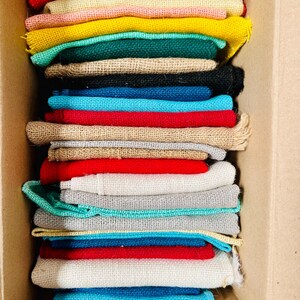 May include: A stack of colorful burlap fabric squares in a cardboard box. The squares are in various colors, including red, yellow, green, blue, and natural burlap. The fabric appears to be folded and stacked neatly.