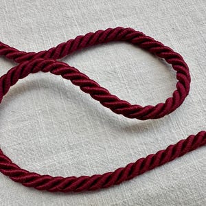 May include: A deep burgundy twisted cord trim featuring a multi-strand braided construction. The cord is arranged in a loose, curved loop, showcasing the consistent spiral texture and the rich, dark red color of the twisted fibers throughout.