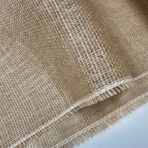 Op de afbeelding: Close-up van een rol natuurlijk jute weefsel. De stof heeft een grove, open weefsel met een lichtbruine kleur. De randen zijn afgewerkt met een wit gestikte rand en een franje.
