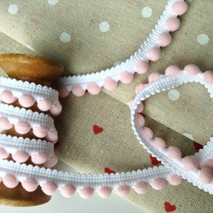 May include: A spool of white trim with pink pom poms. The trim is wrapped around a wooden spool and is lying on a fabric background with white polka dots.