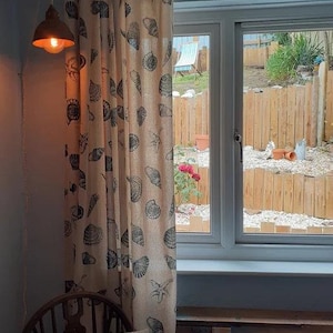 May include: A beige curtain with a seashell pattern hangs in front of a window with a view of a backyard. The curtain has a brown and white pattern of seashells and starfish. The window is open and the view outside is of a wooden fence and a green lawn.