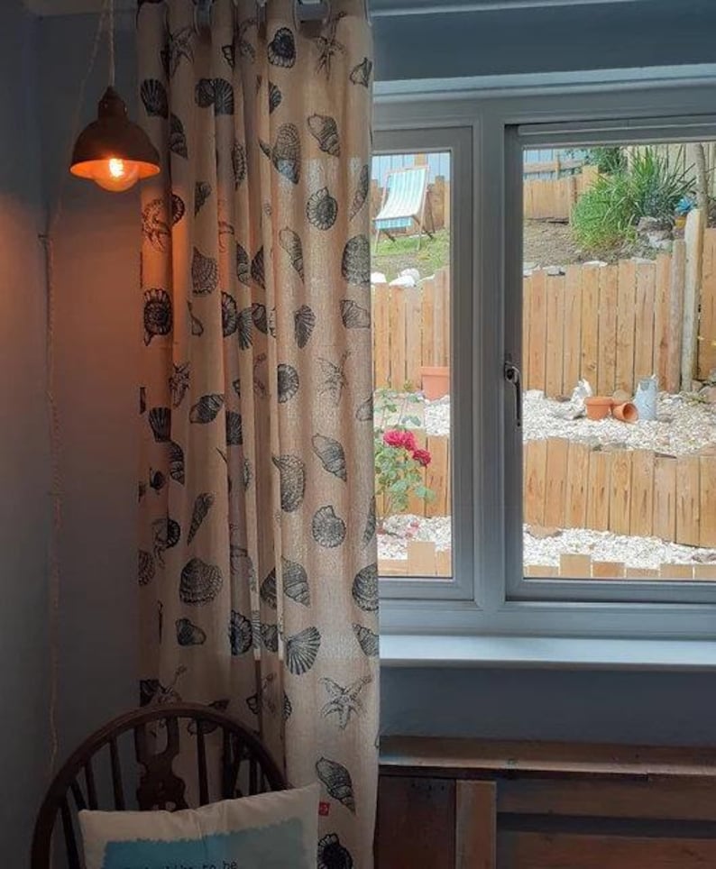 May include: A beige curtain with a seashell pattern hangs in front of a window with a view of a backyard. The curtain has a brown and white pattern of seashells and starfish. The window is open and the view outside is of a wooden fence and a green lawn.