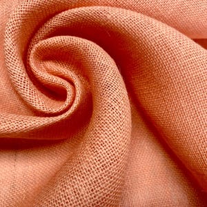 Könnte beinhalten: Nahaufnahme von pfirsichfarbenem Jute-Stoff, eng gewebt mit einem sichtbaren Gittermuster. Der Stoff ist spiralförmig angeordnet und zeigt seine Textur und Farbvariationen. Die Falten erzeugen Schatten, die die Tiefe des Materials hervorheben.