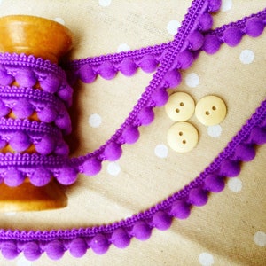 May include: A wooden spool holds a roll of vibrant purple pom-pom trim. Three strands of the trim are visible, along with three small, light-coloured wooden buttons. The trim and buttons are on a beige fabric with white polka dots.