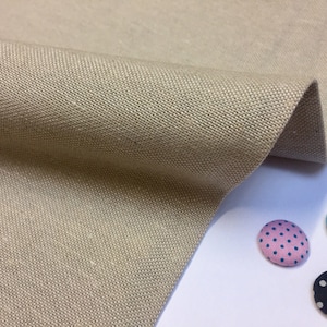 Può includere: Un primo piano di un tessuto di lino beige con una trama testurizzata. Tre bottoni rivestiti in tessuto con pois sono visibili nell'angolo in basso a destra dell'immagine. I bottoni sono rosa con pois blu scuro, blu con pois rosa e nero con pois bianchi.