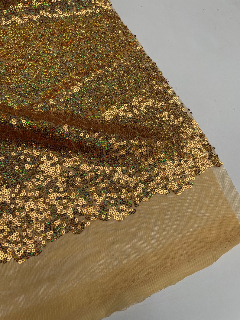 Pode incluir: Close-up de um tecido de lantejoulas douradas. As lantejoulas s&atilde;o densamente compactadas e refletem a luz, criando um efeito brilhante. O tecido &eacute; sobreposto a um suporte de malha bege transparente, adicionando profundidade. Ideal para costura.