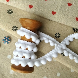 May include: White pom-pom trim wound around a wooden spool, with two buttons in the background. The trim is a popular embellishment for clothing and crafts.