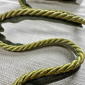 May include: A close-up of a green and gold rope trim with a textured surface. The rope trim is made of two different materials, one is a smooth, shiny fabric and the other is a rougher, textured fabric.
