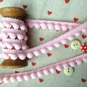 May include: Pink pom-pom trim wound on a wooden spool. The trim is a soft, fluffy pink and is ideal for adding a touch of whimsy to any project.