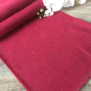 Könnte beinhalten: Ein roter Tischläufer aus Jute mit leicht ausgefransten Kanten. Der Läufer ist in der Mitte gefaltet und hat eine dezente Textur.