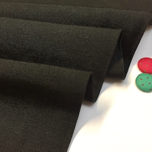 May include: Black fabric with a textured weave. The fabric is folded in thirds and is lying on a white surface. There are three small buttons with polka dots on the surface, one red, one green, and one pink.