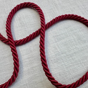 May include: A deep burgundy twisted cord with a smooth, glossy finish. The rope features a consistent spiral texture throughout its length, forming a series of gentle curves and loops that highlight the rich, dark red color of the material.
