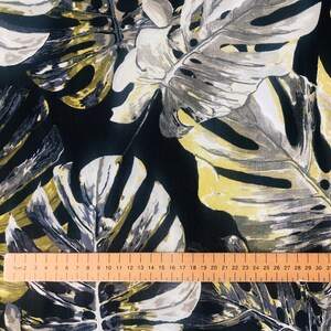 May include: Black fabric with a large-scale tropical leaf print in shades of gray, white, and yellow. A wooden ruler is placed on the fabric, showing measurements in centimeters.