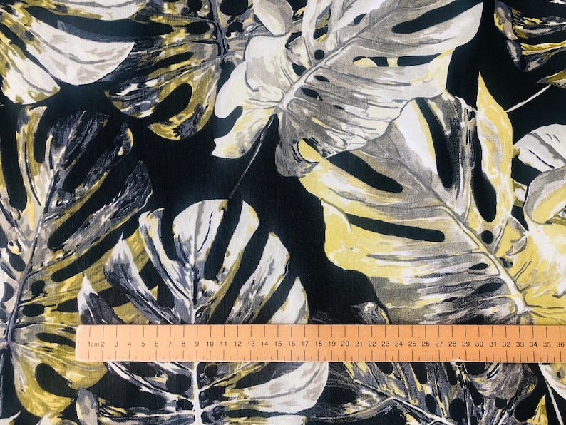 May include: Black fabric with a large-scale tropical leaf print in shades of gray, white, and yellow. A wooden ruler is placed on the fabric, showing measurements in centimeters.