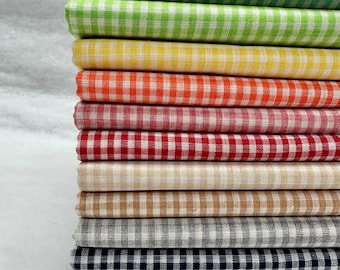 Könnte beinhalten: Ein Stapel gefalteter Stoffe in verschiedenen Farben, jeweils mit einem weiß-farbigen Vichy-Muster. Zu den Farben gehören Grün, Gelb, Orange, Rot, Beige, Grau, Marineblau und Schwarz. Der Stoff ist eng gefaltet und zeigt die Textur des Materials.