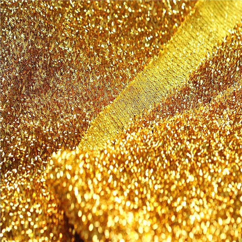 May include: Close-up of a gold-colored fabric with a textured, sparkly surface. The fabric is folded over, creating a layered effect.