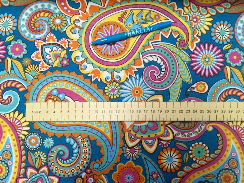 May include: A blue fabric with a paisley pattern in shades of orange, yellow, pink, and green. A ruler is placed on the fabric, with the numbers 1 through 30 visible. The text "BARCLAY" is visible on a blue pen lying on the fabric.