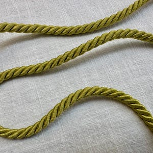May include: A single length of olive green twisted cord trim. The cord features a tightly wound, multi-strand construction with a smooth, slightly lustrous finish, creating a consistent spiral pattern along its entire length.