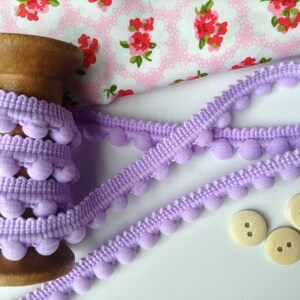 May include: A spool of lavender pom-pom trim with a wooden bobbin. The trim is on a white surface with three wooden buttons.