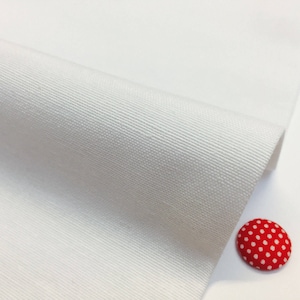 Può includere: Tessuto di cotone bianco con una texture sottile. Il tessuto è piegato a metà e il bordo è visibile. Ci sono tre piccoli bottoni con motivi a pois nell'angolo in basso a destra dell'immagine.